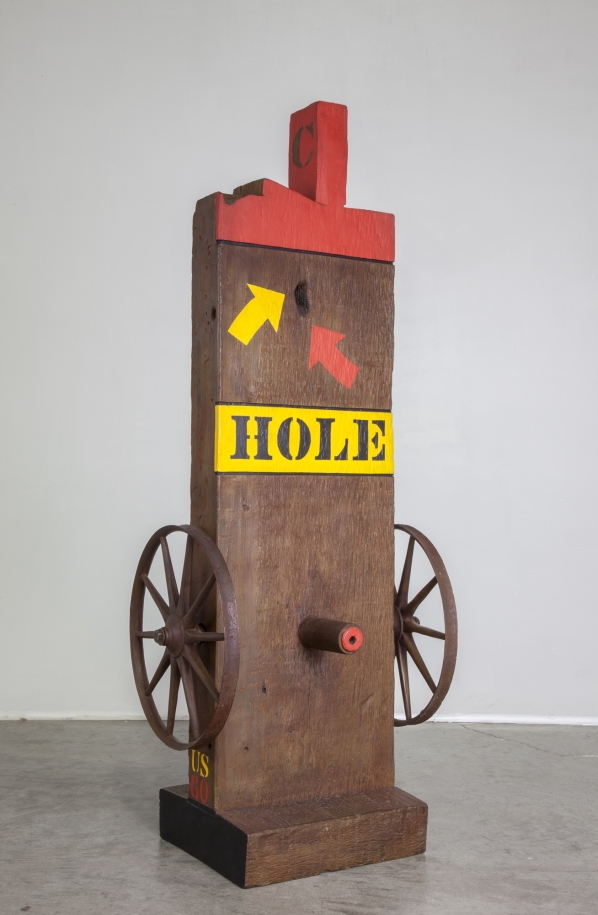 A brown painted bronze sculpture of a beam with a haunched on a base. Wheels have been affixed to the lower right and left of the sculpture, in between the wheels, on the front of the sculpture, is a peg. The work's title, Hole, appears above the wheels, painted in black stenciled letters against a yellow band of paint. Above the title are a yellow and a red arrow, each pointing towards a hole in the sculpture. The tenon and top front of the beam are painted red.