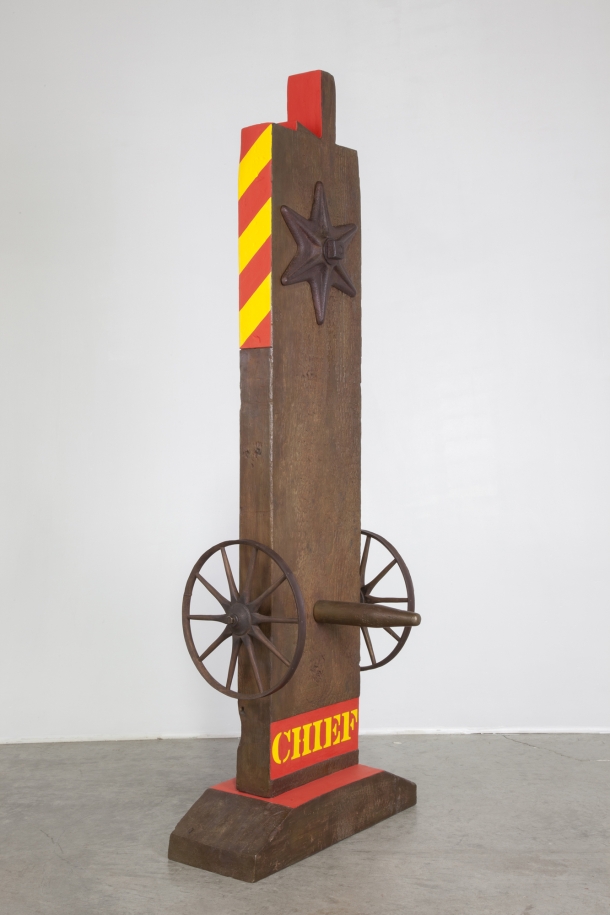 A 63 13/16 by 23 1/8 by 9 1/8 inch painted bronze sculpture of a beam with a haunched tenon, and on a base. The sculpture's title, "Chief," is painted in yellow stenciled letters against a red rectangular ground across the bottom of the work. Above the title a wheel is attached to the right and left sides of the sculpture, and a peg has been placed in the front of the sculpture, in between the wheels. At the top of the sculpture is a six pointed star. The top third of the right and sides of the sculpture are painted with diagonal red and yellow stripes, and the sides left and right sides of the tenon are painted red.