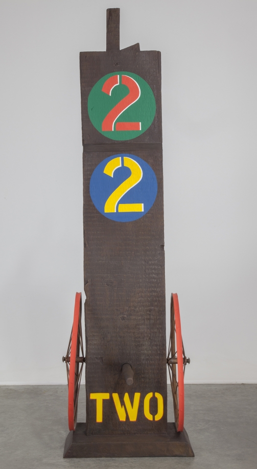 A 61 1/8 by 18 1/2 by 19 1/2 inch painted bronze sculpture of a beam with a haunched tenon, on a base. The work's title, "Two," is painted in yellow stenciled letters across the bottom front of the sculpture. A wheel is attached to the bottom left and right sides of the sculpture, and a peg has been affixed to the front of the sculpture, in between the wheels. At the top of the sculpture is a green circle with a red numeral two, and below that is a blue circle with a yellow numeral two.