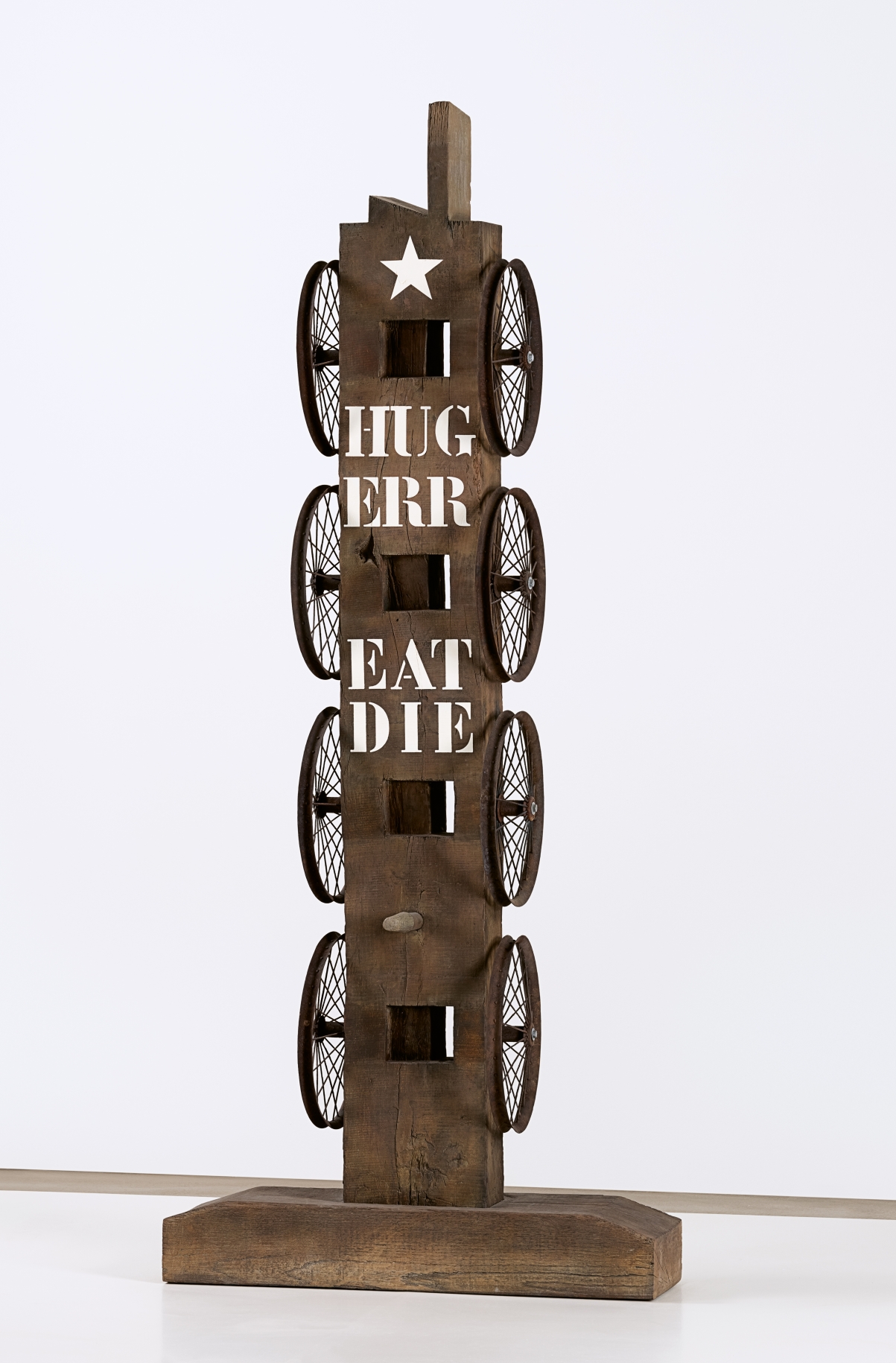 A 83 7/8 by 35 1/2 by 11 13/16 inch painted bronze sculpture consisting of a beam with a haunched tenon on a base. Four wheels run down the left and right sides of the sculpture. At the front top is a white star, below it the words "Hug," "Err," "Eat," and "Die" are painted in white stenciled letters.