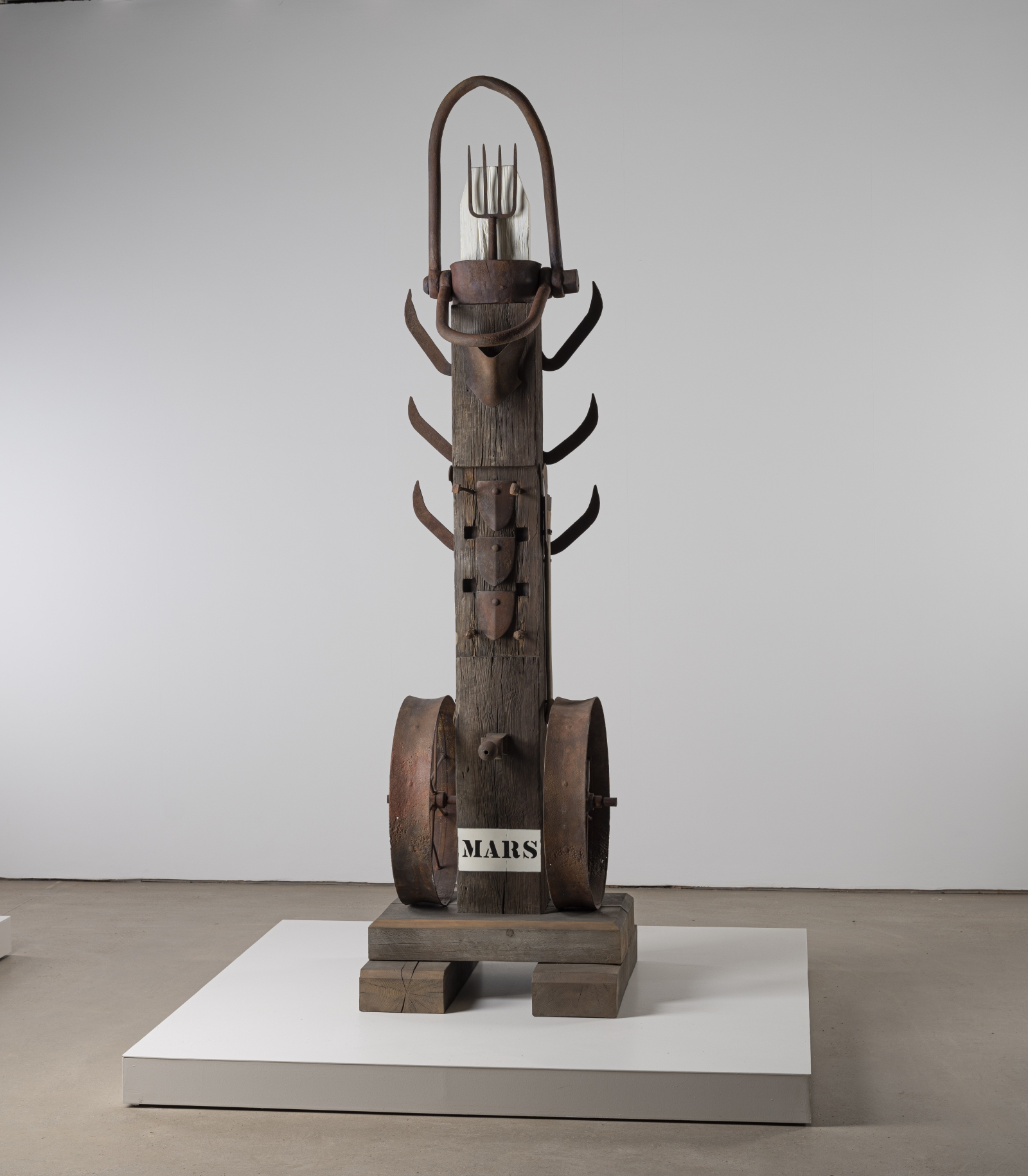 A 131 5/8 by 35 7/8 by 35 7/8 inch painted bronze sculpture consisting of a beam atop a base. The work's title, "Mars," appears in stenciled letters against a white ground across the front bottom of the sculpture. A wheel is affixed to both the bottom right and left sides of the sculpture. Three brush axes have been affixed to both the upper right and upper left sides of, and a pitchfork has been affixed at the top of the sculpture.
