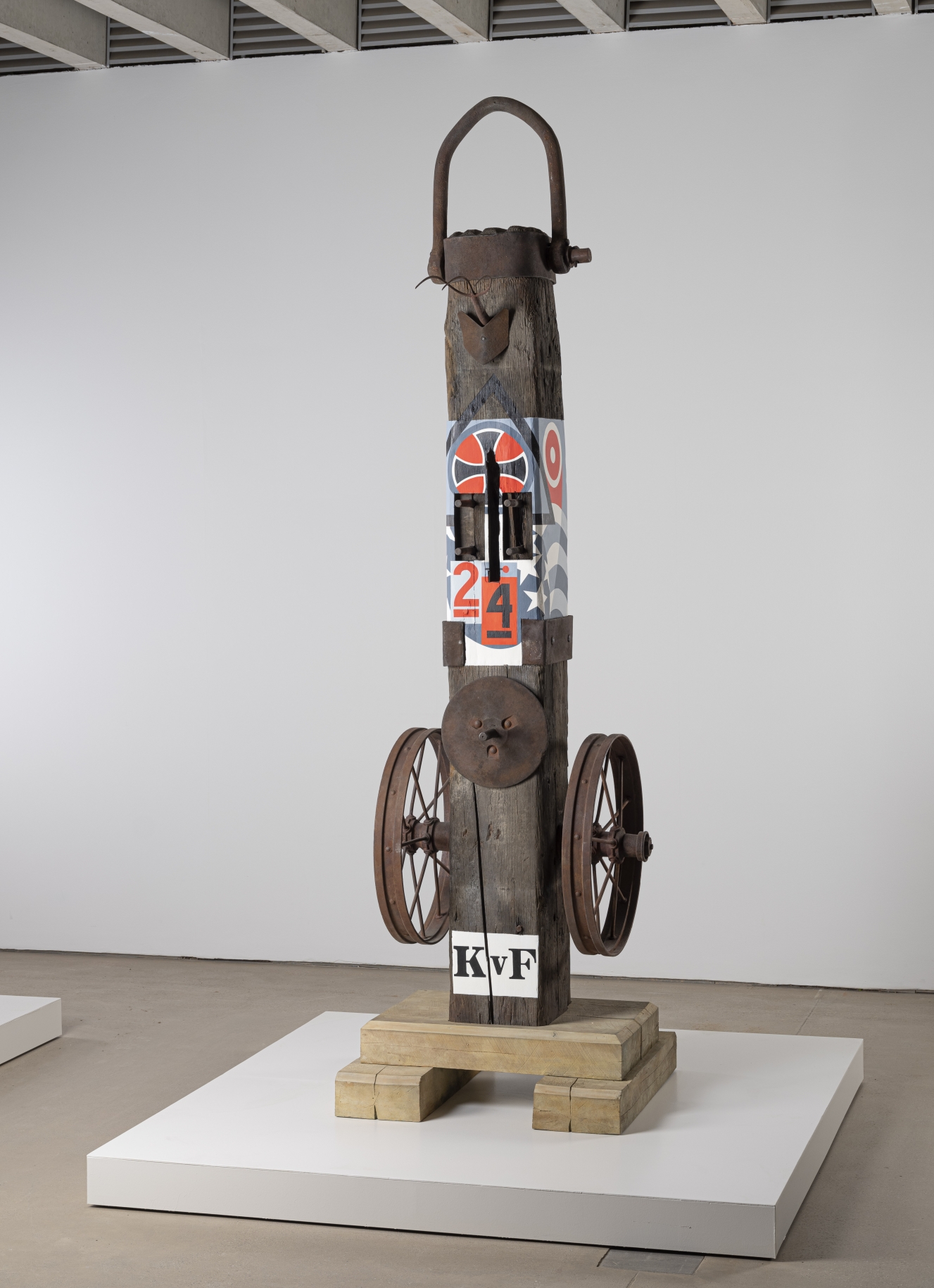 A 138 1/8 by 38 9/16 by 45 1/16 inch painted bronze sculpture consisting of a beam on a base. The work's title, "KvF" appears in stenciled letters against a white ground. A wheel is affixed to the right and left sides of the sculpture. On the front of the sculpture, level with the top of the wheels, is a disk. Above this a section of the sculpture has been painted with red, blue, white, and black designs that include a cross, stars, and the numbers 2 and 4. At the top of the sculpture is a cap with a handle.