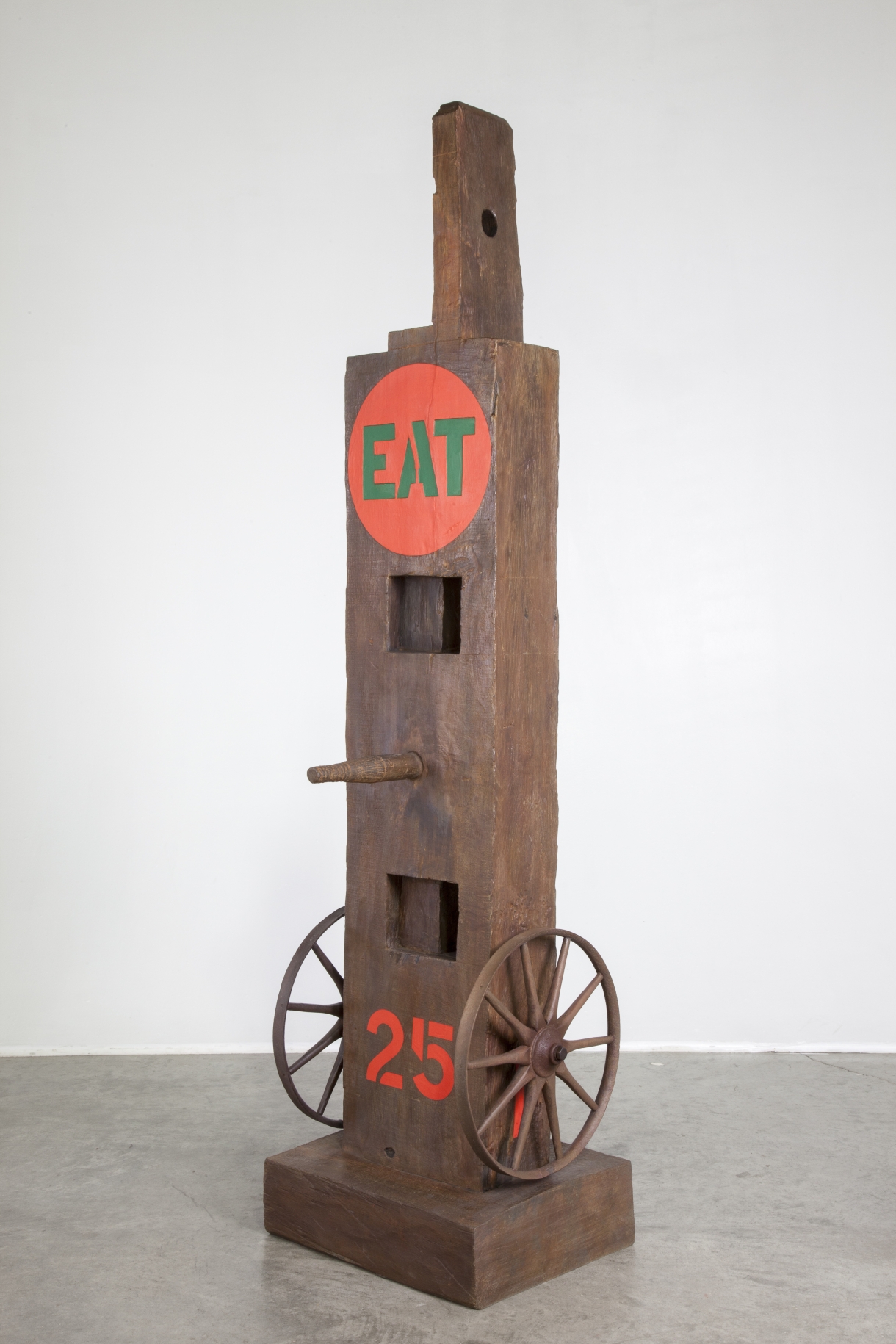 Eat is a 58 5/8 by 15 by 12 5/8 painted bronze sculpture of a beam with a tenon, standing on a base. A wheel is affixed to the right and left sides at the bottom of the sculpture. In between a red number 25 has been painted. A peg protrudes from the center of the sculpture, and at the top of the sculpture is a red circle containing the word "eat" painted in green stenciled letters.