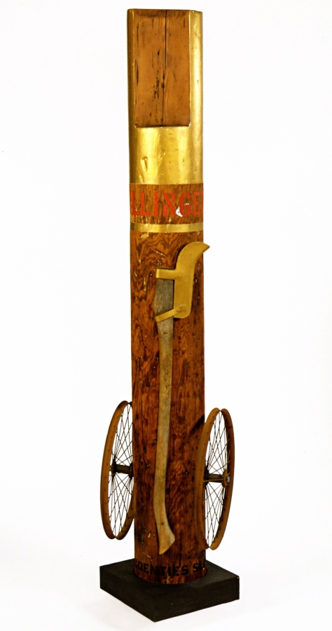 A 75 1/2 × 22 1/2 × 21 inch column with an iron wheel affixed to the right and left lower sides, and standing on a wooden base. An iron axe is attached to the column. The top of the column is painted gold, and below is the work's title, "Dillinger," in red letters. Below the title a gold strip wraps around the column.