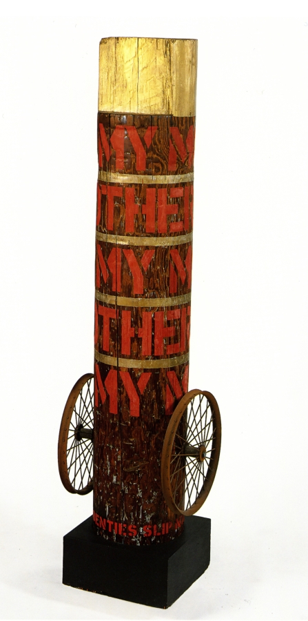 Column with its title "My Mother" painted in five rows around the column in red stenciled letters. The top of the column is painted gold, and thin gold strips around the column separate each row of text. At the bottom of the column, on both sides, is a wheel. 
