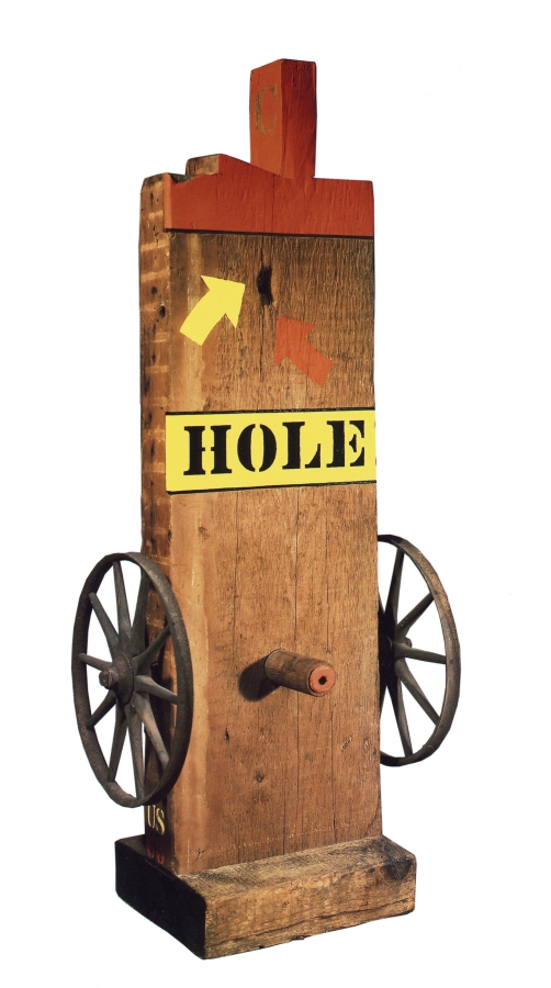 A 45 by 19 by 13 inch sculpture consisting of a wooden beam on a wooden base. Iron and wooden wheels have been affixed to the lower right and left of the sculpture, in between the wheels, on the front of the sculpture, is a wooden peg. The work's title, Hole, appears above the wheels, painted in black stenciled letters against a yellow band of paint. Above the title are a yellow and a red arrow, each pointing towards a hole in the sculpture. The tenon and top front of the beam are painted red.