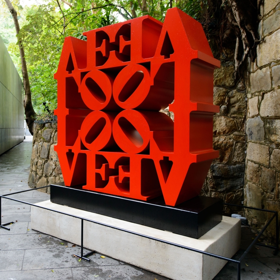 LOVE Wall is a 72 by 72 by 24 inch red polychrome aluminum sculpture consisting of four LOVEs, each with the letters L and a tilted O above the letters V and E. , Two of the LOVEs are upright and two inverted, all facing inward with the four “O”s meeting in the center.