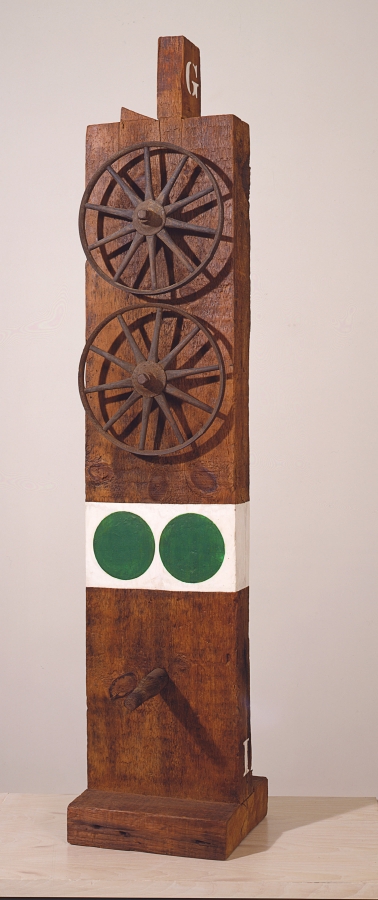 Ge is a wooden sculpture measuring 58 1/2 by 12 by 14 inches that consists of a wooden beam with a tenon on a wooden base. On the top front of the sculpture are two iron and wooden wheels. Below the wheels is a white band of paint with two green circles. Halfway below the white band and the base a wooden peg protrudes from the sculpture.