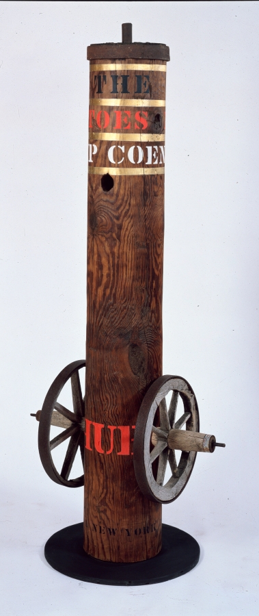 A 83 1/2 by 33 by 26 inch column with its title, "Hub" painted in red stenciled letters towards the bottom of the work, with an iron and wooden wheel affixed to the right and left sides. The work is topped by an iron cap, below are three rows of text separated by a gold band. The top row reads "City of the" in black letters, below it is "Manhattoes" in red, and below that "Coenties Slip" in white letters.